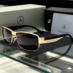 May include: Gold-tone metal sunglasses with dark brown gradient lenses and black temples. The bridge features a small Mercedes-Benz logo, and the temples are engraved with the Mercedes-Benz brand name. Includes a branded box and card.
