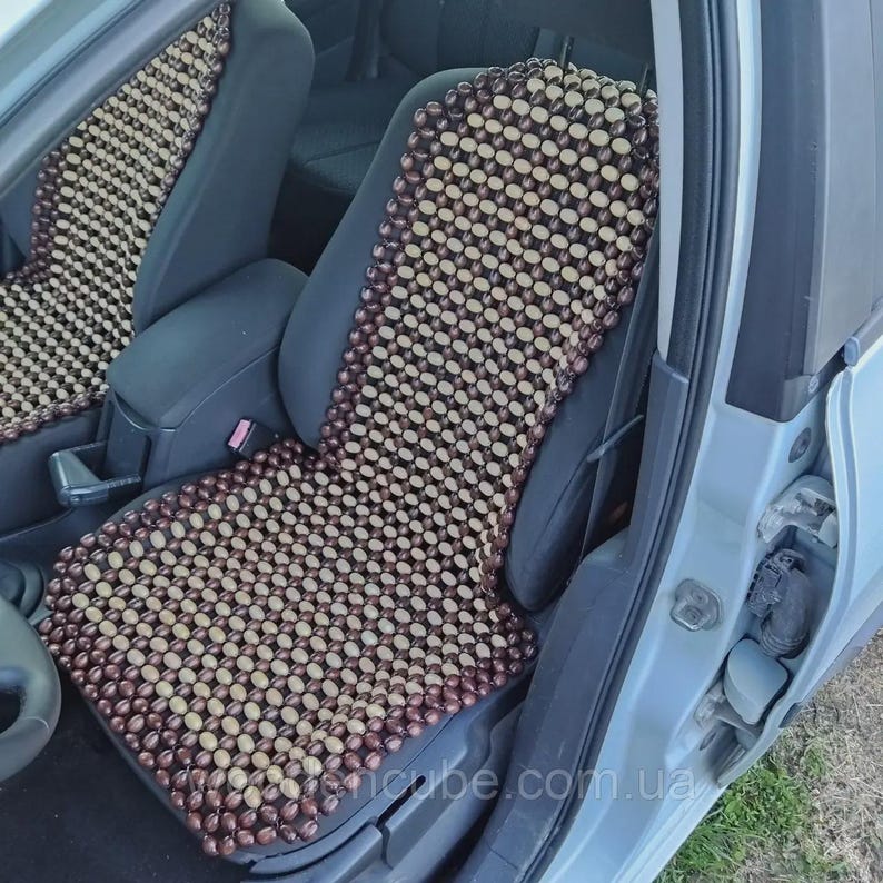 May include: A car seat cover constructed from a grid of smooth, oval-shaped wooden beads in alternating shades of dark brown and light tan. The cover is draped over the front passenger seat of a vehicle, featuring a consistent pattern of beads.