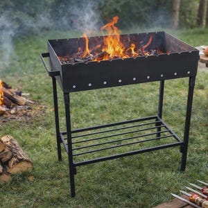 May include: A black metal barbecue grill with a fire burning inside. The grill is on a grassy surface, with a stack of firewood and a picnic table in the background. Skewered food is on a wooden surface.