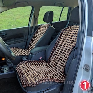 May include: Two car seat covers constructed from small, polished wooden beads arranged in a grid pattern. The beads alternate between dark brown and light tan colors, creating a striped design that covers the backrest and seat of the vehicle chairs.