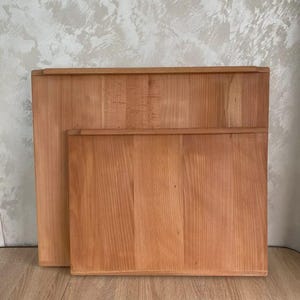 May include: Two rectangular wooden cutting boards with a natural light brown grain pattern. The boards are stacked, with the larger one positioned behind the smaller one. Both boards feature a flat surface and a raised edge along the top side.