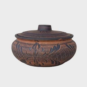 Könnte beinhalten: Braune Keramikschüssel mit Deckel, verziert mit einem geschnitzten Blumenmuster. Die Schüssel hat dunkelblaue Akzente und eine strukturierte Oberfläche. Der Deckel hat einen abgerundeten Griff. Durchmesser ca. 20 cm, dekoratives Stück.