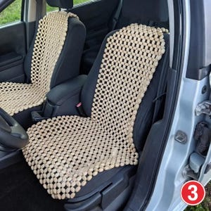May include: Two beige wooden beaded seat covers installed on the front seats of a vehicle. The covers feature a grid of small, oval-shaped light-colored wooden beads connected by string, covering the backrest and seat cushion of each car seat.