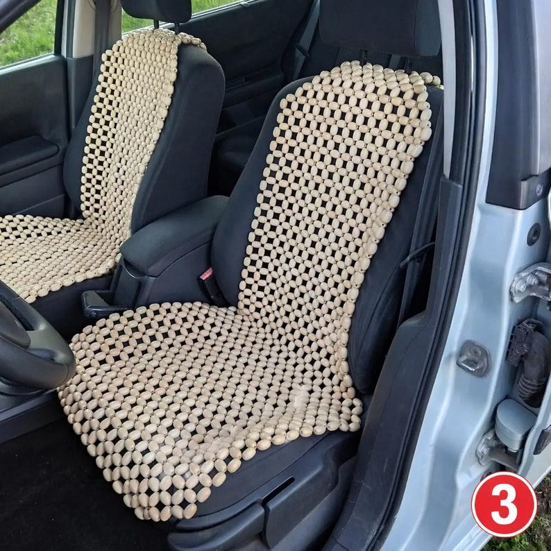 May include: Two beige wooden beaded seat covers installed on the front seats of a vehicle. The covers feature a grid of small, oval-shaped light-colored wooden beads connected by string, covering the backrest and seat cushion of each car seat.