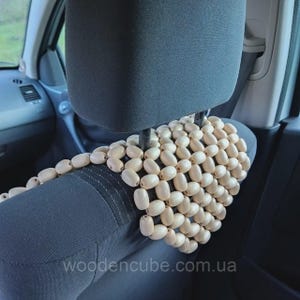 May include: A car seat cover constructed from numerous light-colored, smooth, oval-shaped wooden beads strung together in a grid pattern. The beaded cover is draped over the dark gray fabric headrest and upper back section of a vehicle seat.