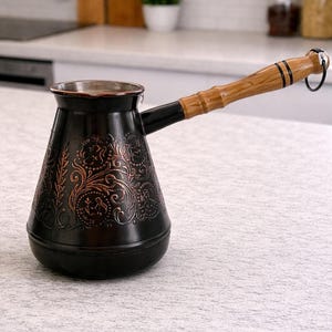 Op de afbeelding: Een zwarte en koperkleurige Turkse koffiepot met een sierlijk bloemmotief. De pot heeft een lange, houten handgreep en een zwarte tuit. De pot staat op een wit oppervlak in een keuken.