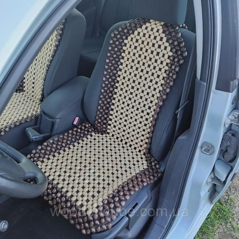 May include: A car seat cover constructed from numerous small, oval-shaped wooden beads. The design features a central panel of light beige beads surrounded by a border of dark brown beads, all connected by a grid pattern to fit the seat shape.