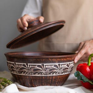 Könnte beinhalten: Eine große, braune Keramikschüssel mit Deckel, mit einem dekorativen weißen Blumenband in der Mitte. Die Schüssel ist offen und zeigt ihr Inneres. Der Deckel wird angehoben und zeigt das Design und die Handwerkskunst der Schüssel.