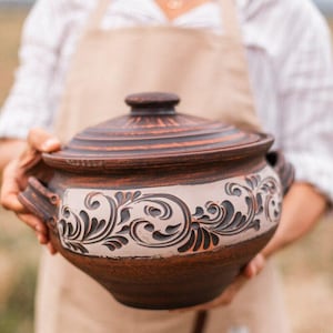 Könnte beinhalten: Ein brauner Keramikkochtopf mit Deckel und einem dekorativen Blumendesign in einer helleren Farbe. Der Topf hat zwei Griffe und wird von einer Person mit einer Schürze gehalten. Der Topf wird wahrscheinlich zum Kochen und Servieren von Speisen verwendet.