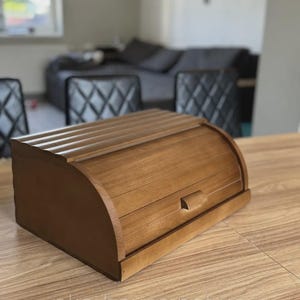 Könnte beinhalten: Ein mittelbrauner Brotkasten aus Holz mit einem gewölbten, lamellierten Rolldeckel und einem kleinen rechteckigen Griff. Der Kasten hat eine glatte, polierte Oberfläche und steht auf einer hellen Holzmaserung. Der Text eko-houss.net.ua ist sichtbar.