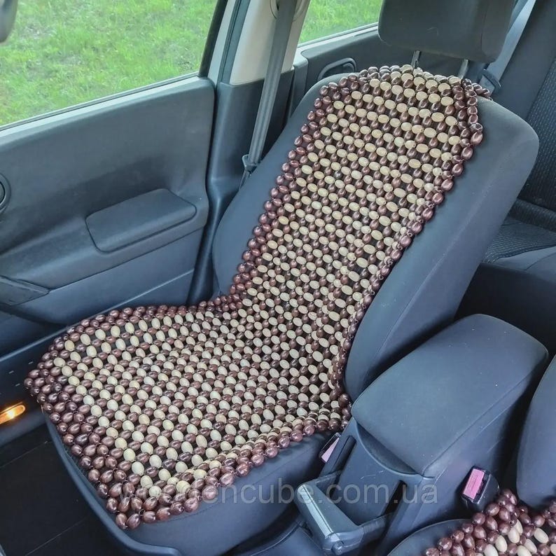May include: A car seat cover constructed from a grid of alternating light beige and dark brown wooden beads. The cover is draped over the front passenger seat of a vehicle, featuring a rectangular shape that extends from the headrest to the seat base.
