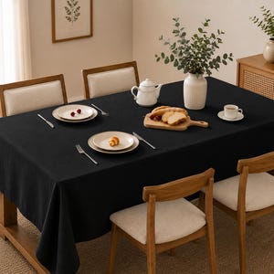 May include: A rectangular black tablecloth draped over a wooden dining table. The table is set with white ceramic plates, silver forks and knives, a white teapot, a wooden serving board with sliced bread, and a white cup and saucer.