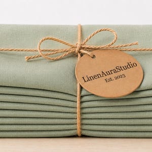 May include: A stack of sage green fabric napkins tied with thin brown twine in a bow. A round, tan-coloured paper tag is attached to the twine, featuring the printed text "LinenAuraStudio" and "Est. 2023" in a dark, serif font centred on the tag.