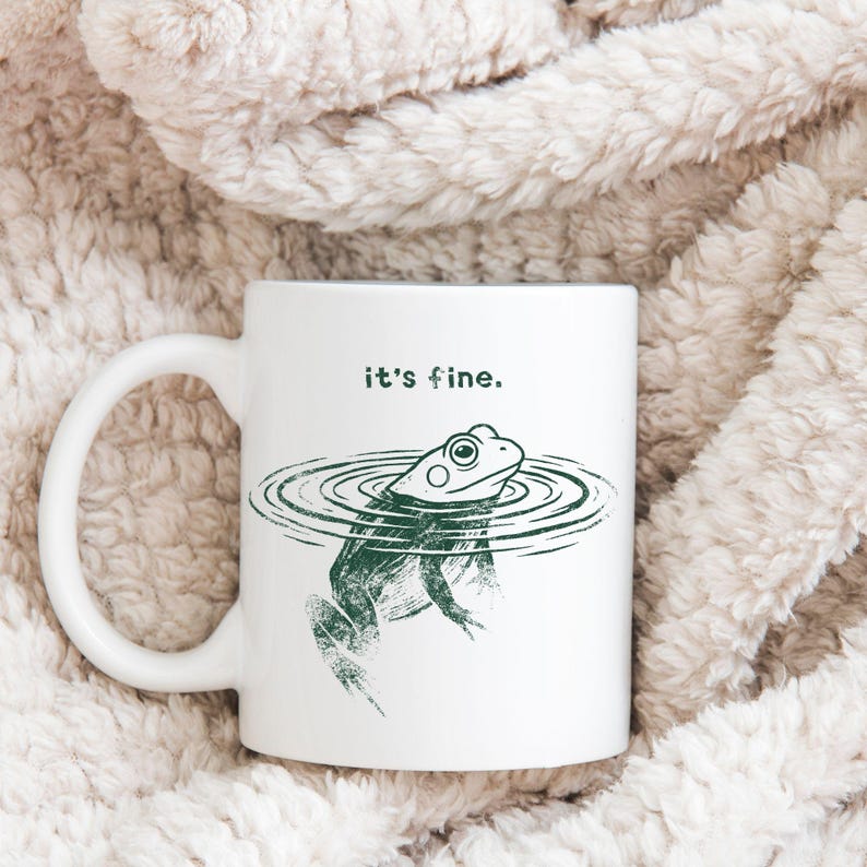 May include: White ceramic mug featuring a green illustration of a frog in water with the text "it's fine." The mug has a large handle and is set against a soft, textured background.