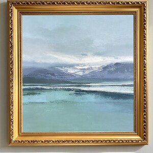 Op de afbeelding: Een vierkant landschapsschilderij in een decoratieve gouden lijst met een serene scène van kalm blauwgroen water op de voorgrond, met verre paarsgetinte bergen onder een zachte, bewolkte lucht. De tekst LUCKYARTSGALLERY staat onderaan.