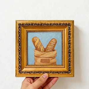 Peut inclure: Une peinture à l'huile dans un cadre carré doré orné représentant deux baguettes dorées sortant d'un sac en papier beige sur un fond bleu clair. L'œuvre présente des coups de pinceau visibles et une texture en empâtement.