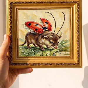 Può includere: Un dipinto a olio quadrato in una cornice dorata decorativa raffigurante una piccola creatura marrone con due piccole corna e ali di coccinella rosse con punti neri. La creatura si trova su una grande foglia verde coperta di gocce d'acqua.