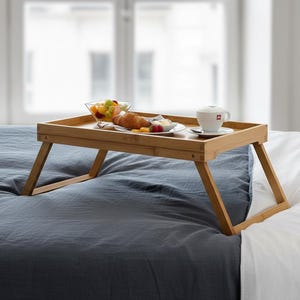 May include: A bamboo breakfast tray with folding legs, set on a bed with a blue comforter and white sheets. The tray holds a cup of coffee, a plate of food, and a bowl of fruit. The tray is approximately 18 inches long.
