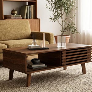 May include: A rectangular dark brown wooden coffee table featuring four tapered legs, an open storage shelf, and a side section with horizontal slats. The top surface holds a clear glass vase with a plant, books, and two cylindrical decorative objects.