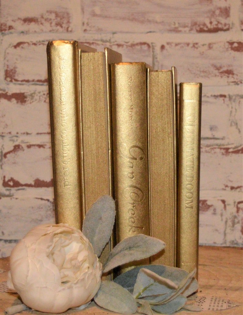 May include: A set of five gold-painted books with titles visible on the spines. The titles are "The Teacher Who Couldn't Read", "Robert Moore", "Inn Creek", and "The Fate of Doom".