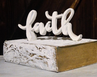 May include: A decorative wooden block with the word "faith" in white script. The block is painted white with gold edges, creating a rustic, vintage aesthetic. The item is likely a home decor piece.
