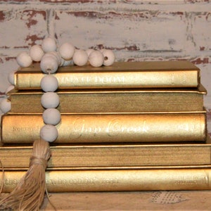 May include: A stack of five gold-painted hardback books with white text on the spines. The books are decorated with a white beaded garland and a brown tassel.