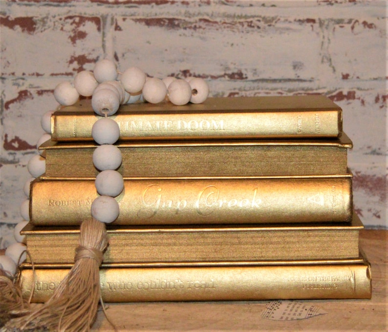 May include: A stack of five gold-painted hardback books with white text on the spines. The books are decorated with a white beaded garland and a brown tassel.