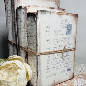 May include: A stack of three rectangular books with distressed white covers featuring black cursive script, postal stamps, and dark brown edges. The books are bound together with a thin piece of brown twine and accompanied by a large white flower.