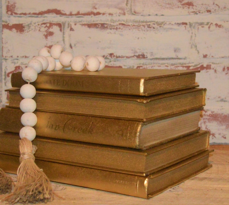 May include: A stack of five gold-painted books with a white bead garland on top. The books have titles such as "Cafe Diem" and "Imp Creek".