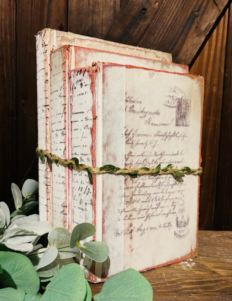 Pode incluir: Uma pilha de tr&ecirc;s livros de estilo vintage com capas brancas e bordas vermelhas. Os livros s&atilde;o decorados com texto escrito &agrave; m&atilde;o e t&ecirc;m um aspecto r&uacute;stico e envelhecido. Os livros est&atilde;o amarrados com barbante e folhas verdes.