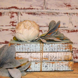 May include: A stack of three vintage books with a light blue painted finish and white text. The books are tied together with twine. A white faux peony flower and gray-green foliage are placed on top of the books.