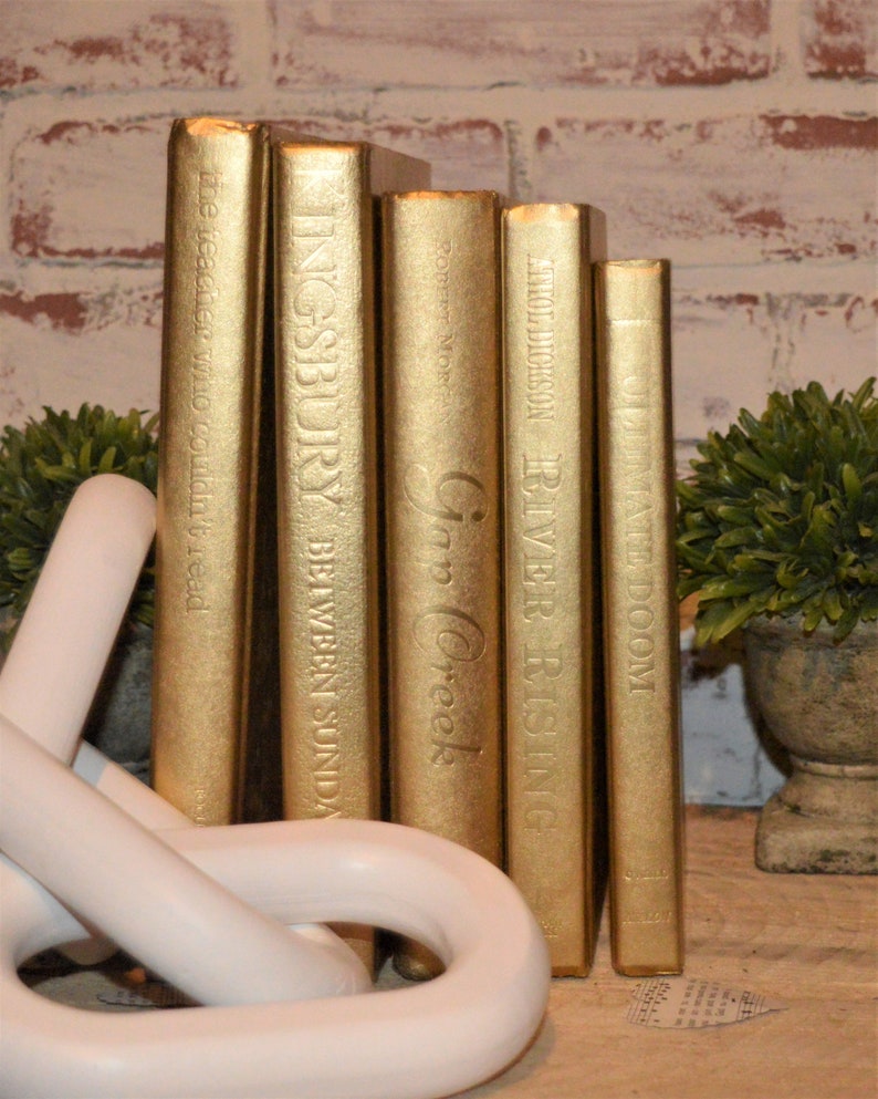 May include: A set of five gold-painted books with titles visible on the spines. The titles are "The Teacher Who Couldn't Read", "Ingsburry Between Sunday", "Gap Creek", "River Rising", and "Ultimate Doom".