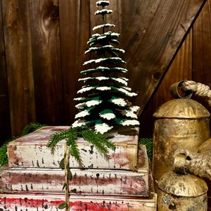 Pode incluir: Uma pequena &aacute;rvore de Natal verde com neve artificial branca, empilhada em cima de tr&ecirc;s livros antigos com p&aacute;ginas vermelhas e brancas, e dois sinos dourados com al&ccedil;as de corda.
