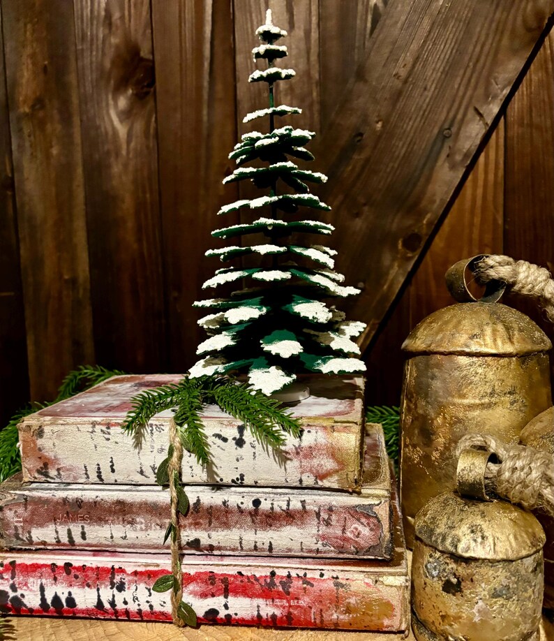 Pode incluir: Uma pequena &aacute;rvore de Natal verde com neve artificial branca, empilhada em cima de tr&ecirc;s livros antigos com p&aacute;ginas vermelhas e brancas, e dois sinos dourados com al&ccedil;as de corda.