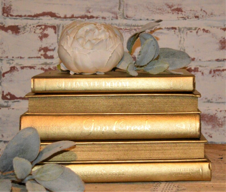 May include: A stack of five gold-painted books with white lettering on the spines. The top book has the title "Ultimate Doom" and the second book has the title "Gap Creek". A white faux flower sits on top of the books.