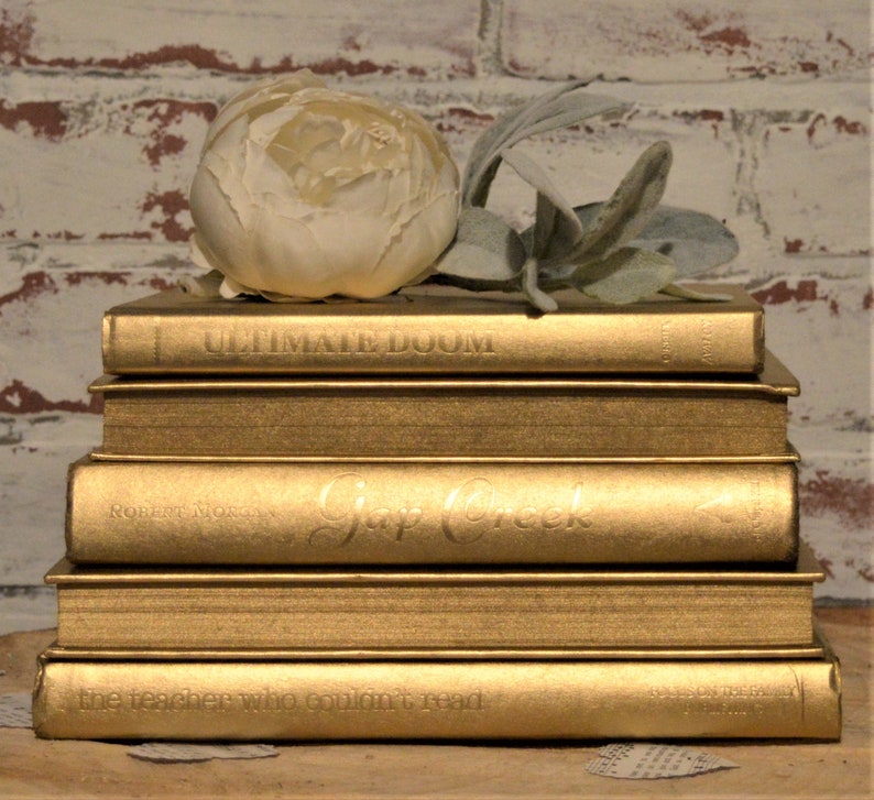 May include: A stack of five gold-coloured hardback books with white text on the spines. The top book has the title "Ultimate Doom" on the spine. The second book has the title "Gap Creek" on the spine. The third book has the title "The Teacher Who Couldn't Read" on the spine. A white flower sits on top of the books.