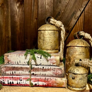 Pode incluir: Tr&ecirc;s sinos de metal pintados de ouro com al&ccedil;as de corda, empilhados em cima de quatro livros de estilo vintage com um design pintado de branco e vermelho.