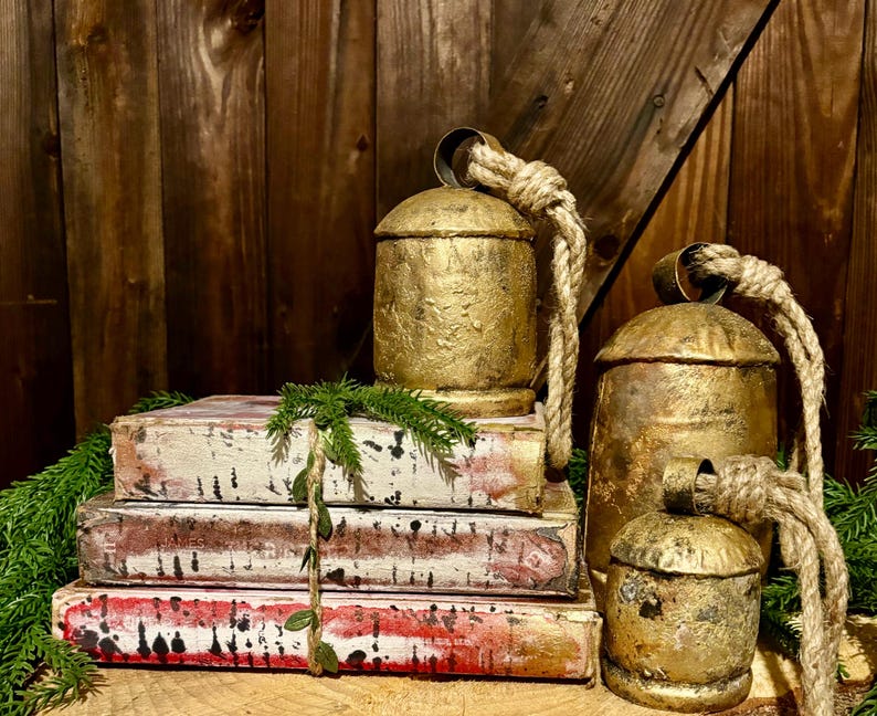 Pode incluir: Tr&ecirc;s sinos de metal pintados de ouro com al&ccedil;as de corda, empilhados em cima de quatro livros de estilo vintage com um design pintado de branco e vermelho.