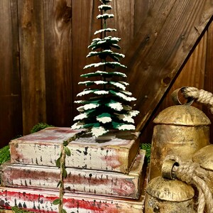 Pode incluir: Uma pequena &aacute;rvore de Natal verde com neve artificial branca em seus galhos est&aacute; em cima de uma pilha de quatro livros antigos. Os livros est&atilde;o amarrados com uma guirlanda verde. Dois sinos dourados com al&ccedil;as de corda est&atilde;o em primeiro plano.