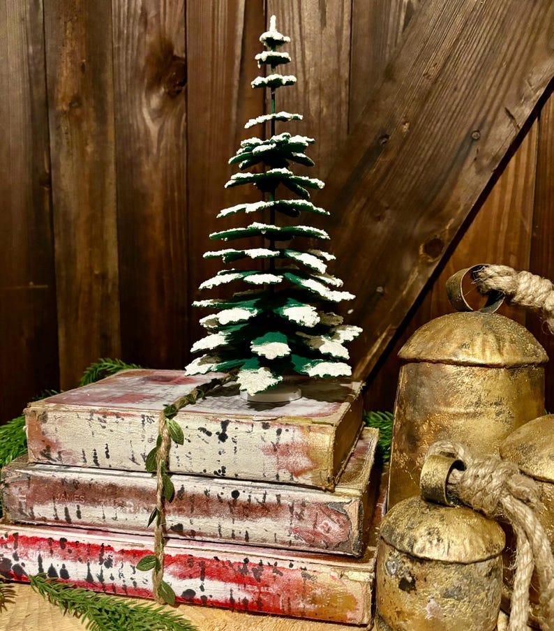 Pode incluir: Uma pequena &aacute;rvore de Natal verde com neve artificial branca em seus galhos est&aacute; em cima de uma pilha de quatro livros antigos. Os livros est&atilde;o amarrados com uma guirlanda verde. Dois sinos dourados com al&ccedil;as de corda est&atilde;o em primeiro plano.