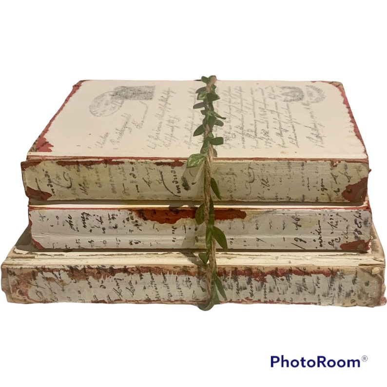 Pode incluir: Uma pilha de tr&ecirc;s livros de estilo vintage com bordas pintadas de vermelho e p&aacute;ginas brancas com texto escrito &agrave; m&atilde;o. Os livros est&atilde;o amarrados com uma guirlanda verde.