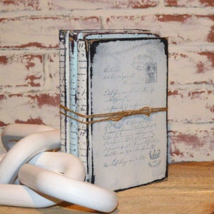 May include: A stack of three vintage-style books with blue and white covers, tied together with twine. The books have a distressed look and are decorated with handwritten text and stamps.