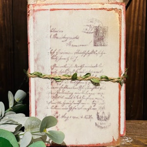 Pode incluir: Uma pilha de di&aacute;rios de estilo vintage com uma capa branca desgastada e bordas vermelhas. Os di&aacute;rios s&atilde;o amarrados com barbante e decorados com folhas verdes.