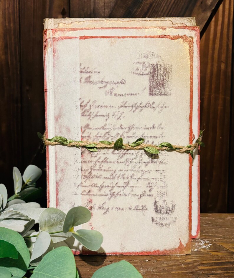 Pode incluir: Uma pilha de di&aacute;rios de estilo vintage com uma capa branca desgastada e bordas vermelhas. Os di&aacute;rios s&atilde;o amarrados com barbante e decorados com folhas verdes.