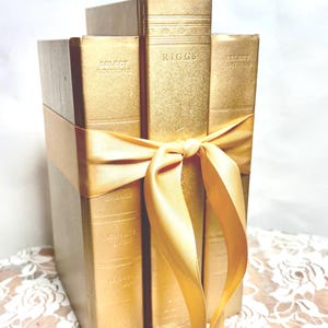 May include: A stack of three books with metallic gold covers tied together with a matching gold satin ribbon. The central book spine features the text RIGGS, while the outer books are labeled SELECT EDITIONS. The books stand upright in a vertical row.
