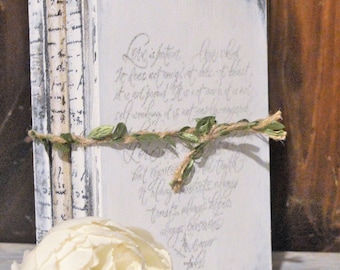 May include: A stack of decorative books with a distressed white finish. The front book cover features elegant script with the words "Love is patient." A twine and leaf garland wraps around the books, and a white flower is in the foreground.