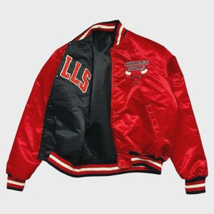 Op de afbeelding: Een rode Chicago Bulls bomberjack van satijn. De jas heeft het teamlogo en de tekst op de voorkant en de letters "LLS" aan de binnenkant. De jas heeft zwart, wit en rood gestreepte boorden aan de manchetten, kraag en zoom.