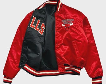 May include: A red satin Chicago Bulls bomber jacket. The jacket features the team logo and text on the front and the letters "LLS" on the inside. The jacket has black, white, and red striped trim on the cuffs, collar, and hem.
