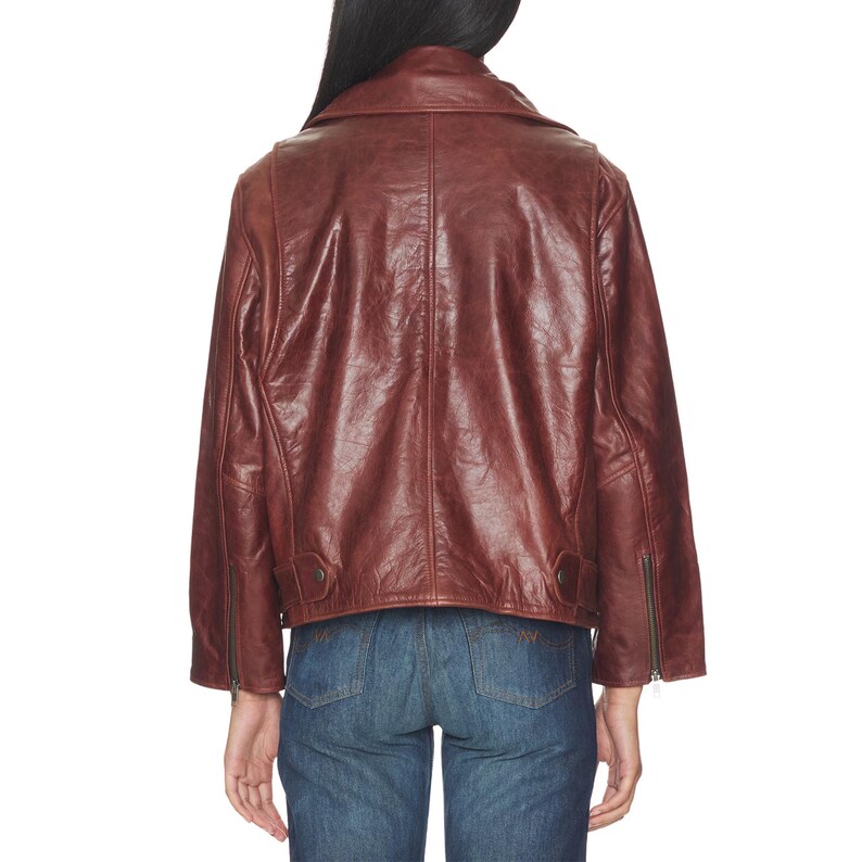 Premium women’s burgundy leather biker jacket featuring an oversized fit, asymmetrical zipper closure, wide lapel collar, vintage finish, adjustable belt detail, and classic moto style paired with denim for a stylish streetwear look.