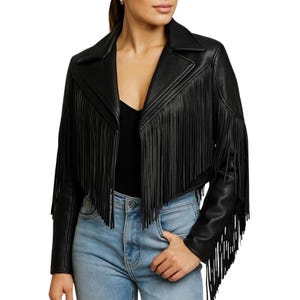 Könnte beinhalten: Schwarze Lederjacke mit langen Fransen. Die Jacke hat einen klassischen Kragen und eine verkürzte Passform. Die Ärmel sind lang und haben ebenfalls Fransen. Die Jacke wird über einem schwarzen Oberteil und einer blauen Jeans getragen.